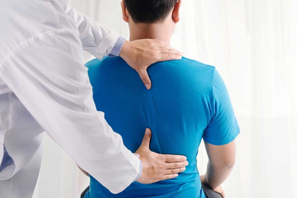 An adult holding their lower back while walking in a park, showing mild discomfort.